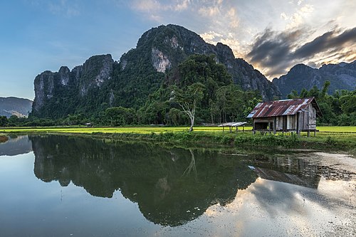 Water reflection of karst mountains, wooden hut, trees and colorful clouds at sunset with green paddy fields, Vang Vieng, Laos